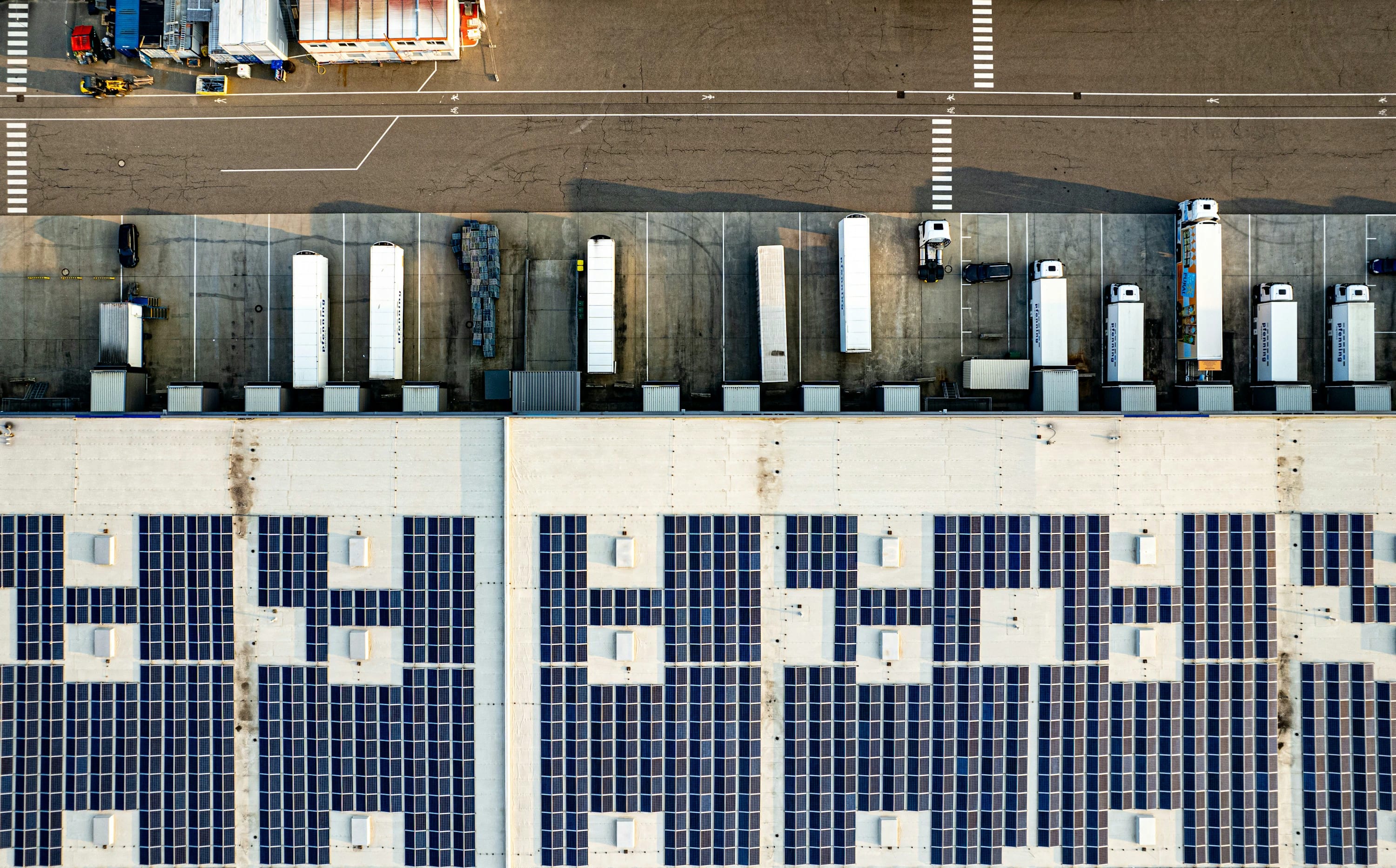 Top-down luchtfoto van een modern logistiek gebouw met laadperrons, vrachtwagens en zonnepanelen.